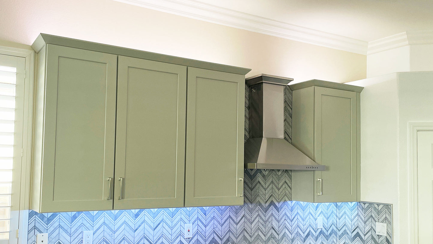 Modern kitchen with green cabinets, a stainless steel range hood, and a herringbone tile backsplash. Under-cabinet lighting highlights the countertop.