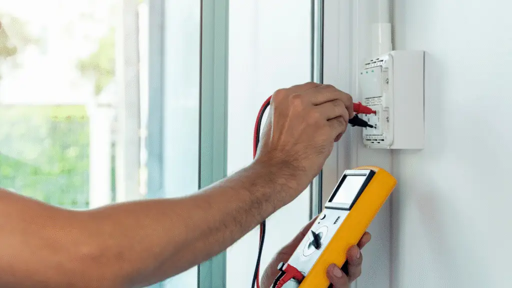 An electrician using a multimeter to test electrical equipment mounted on a wall.