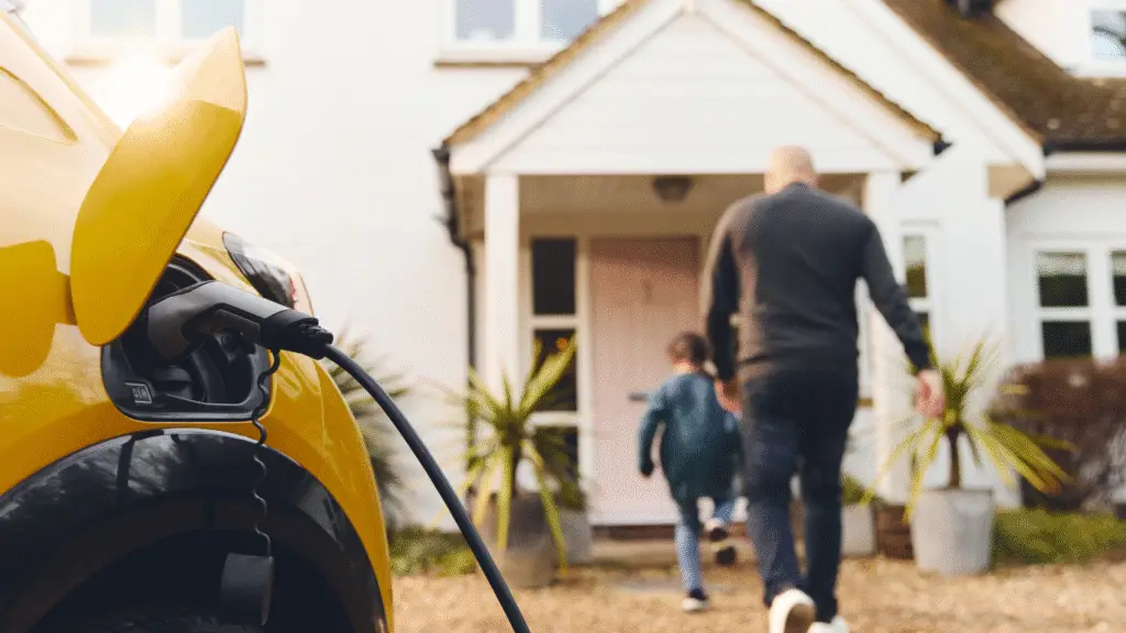 Yellow electric vehicle plugged into a charging station outside a residential home with a man and child approaching the entrance.