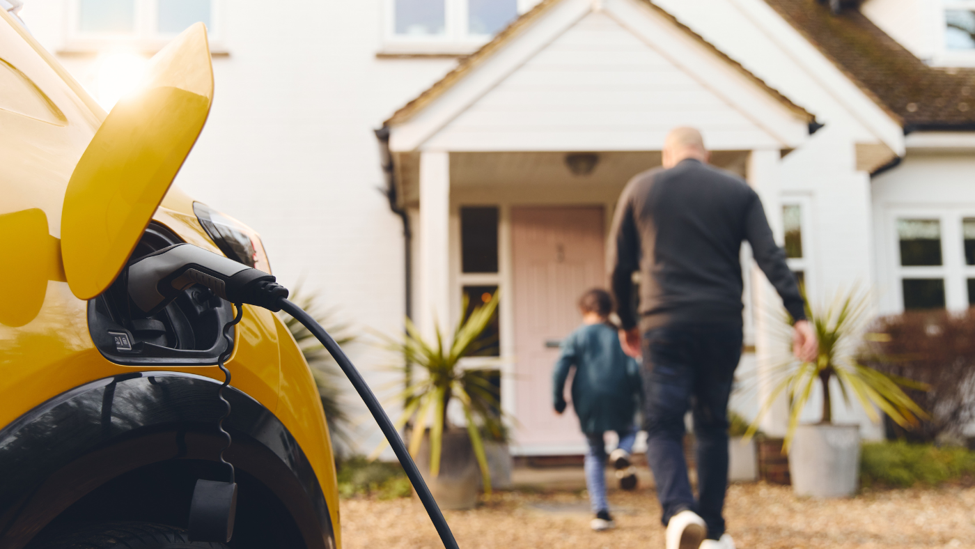 Yellow electric vehicle plugged into a charging station outside a residential home with a man and child approaching the entrance.